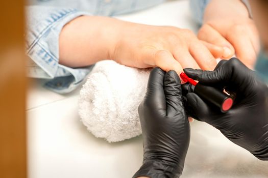 Manicure varnish painting. Close-up of a manicure master wearing rubber black gloves applying red varnish on a female fingernail in the beauty salon