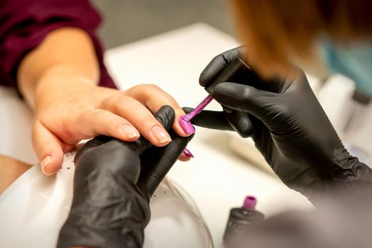 Professional manicure. A manicurist is painting the female nails of a client with purple nail polish in a beauty salon, close up. Beauty industry concept