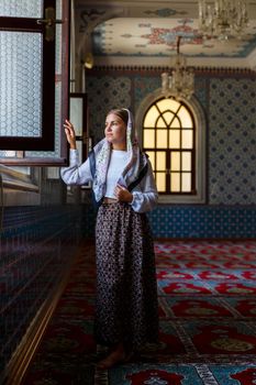 A beautiful Ukrainian woman in a scarf on her head stands in the mosque by the window and prays for peace.