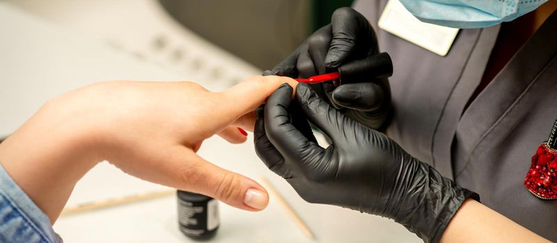 Manicure varnish painting. Close-up of a manicure master wearing rubber black gloves applying red varnish on a female fingernail in the beauty salon