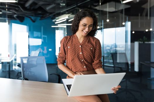 Young beautiful business woman working with laptop smiling and happy, Hispanic woman inside modern office building wearing glasses and curly hair.