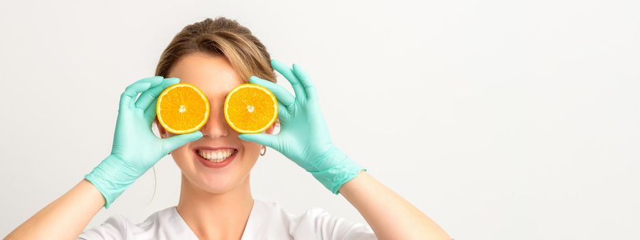 Portrait of young caucasian smiling female beautician covering eye with two orange slices wearing gloves over a white background