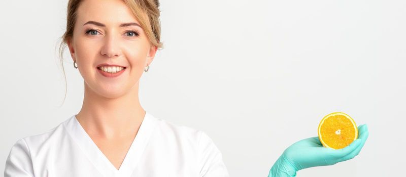 Smiling female nutritionist holding a sliced orange, looking at camera over white background, healthy diet concept