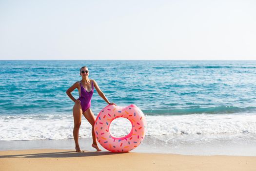 Attractive young woman in a bright swimsuit posing on the beach with an inflatable ring. Beautiful blond woman with long hair relaxing by the ocean. The concept of a sports model, swimwear