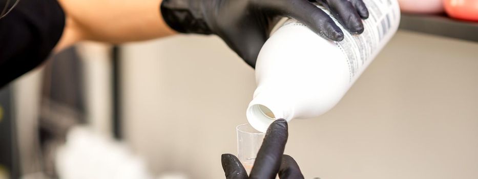 A hairdresser in black gloves is preparing hair dye with a bottle in a hair salon, close up