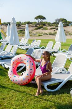A slender young woman holds an inflatable circle in her hands, standing on the green grass. Attractive model with perfect body. Vacation concept, girl goes swimming in the sea