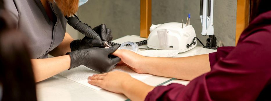 Manicure master wearing protective mask uses electric nail file machine in a nail salon, close up