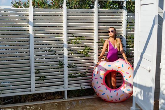 A young woman in a stylish swimsuit and sunglasses holds an inflatable donut ring in her hand. Beautiful happy girl posing and having fun in the sun. Vacation. travel