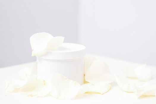 White jar with cream among delicate white rose flowers petals on a light background. Natural organic cosmetics concept, close up, mock-up