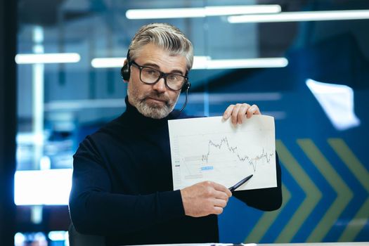 Video call, senior gray-haired businessman looking at camera and showing report financial graph, man not satisfied with financial report, online meeting with colleagues executives investors.