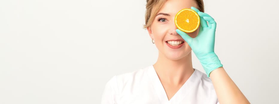 Portrait of young caucasian smiling female beautician covering eye with an orange slice wearing gloves over a white background