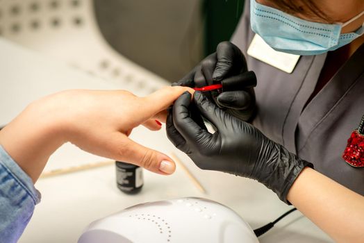 Manicure varnish painting. Close-up of a manicure master wearing rubber black gloves and a protective mask applying red varnish on a female fingernail in the beauty salon