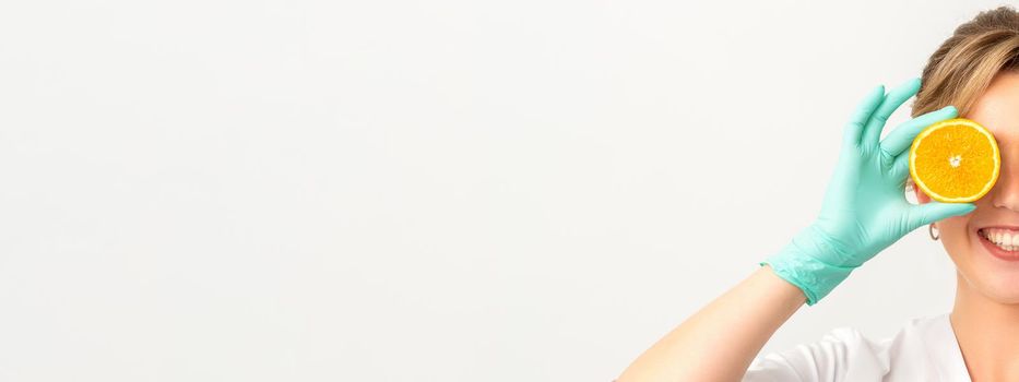 Portrait of young caucasian smiling female beautician covering eye with an orange slice wearing gloves over a white background