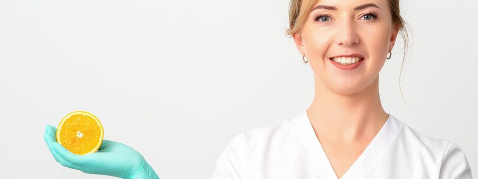 Smiling female nutritionist holding a sliced orange, looking at camera over white background, healthy diet concept