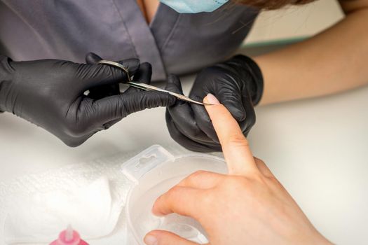 Manicure master removes cuticles from female nails with scissors wearing protective gloves in manicure salon