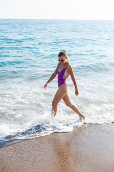 An attractive young woman comes out of the sea in a swimsuit. A girl in a swimsuit walks under the bright summer sun along the sand of the Mediterranean coast. Rest by the sea. Selective focus