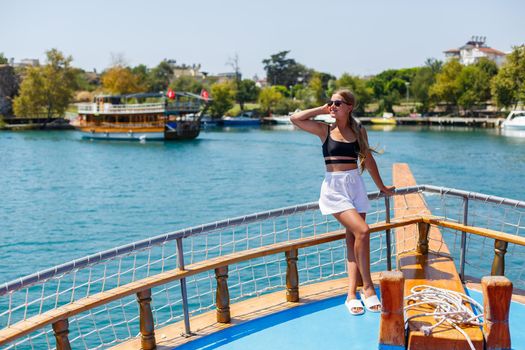 A young slender tanned woman in white shorts and a top, wearing sunglasses. Walk on a hot sunny day on a yacht along the river