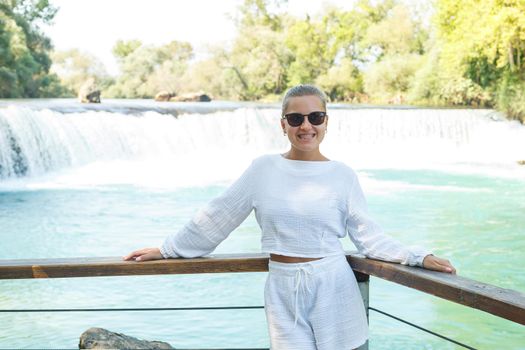 A beautiful young woman in white shorts and a shirt against the backdrop of a mountain waterfall against the backdrop of a dense forest. Beautiful landscape