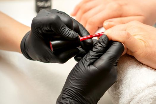 Manicure varnish painting. Close-up of a manicure master wearing rubber black gloves applying red varnish on a female fingernail in the beauty salon