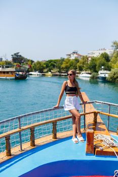 A young slender tanned woman in white shorts and a top, wearing sunglasses. Walk on a hot sunny day on a yacht along the river