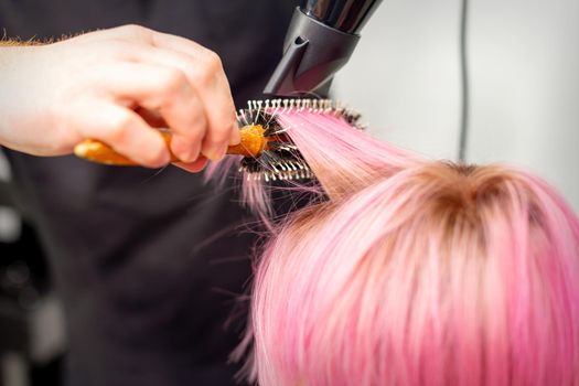 Drying short pink hair of young caucasian woman with a black hairdryer and black round brush by hands of a male hairdresser in a hair salon, close up