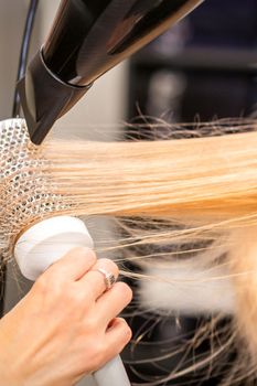 Drying straight blond hair with black hairdryer and white round brush in hairdresser salon, close up