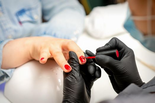 Manicure varnish painting. Close-up of a manicure master wearing rubber black gloves applying red varnish on a female fingernail in the beauty salon