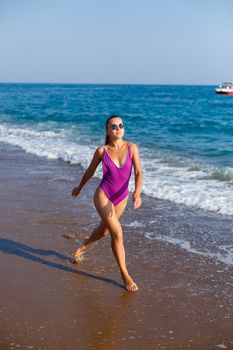 A slender woman in a swimsuit walks along the waves of the ocean on a tropical beach on a sunny day. Summer vacation concept by the sea. Selective focus