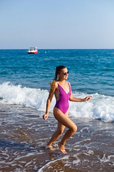 A slender woman in a swimsuit walks along the waves of the ocean on a tropical beach on a sunny day. Summer vacation concept by the sea. Selective focus