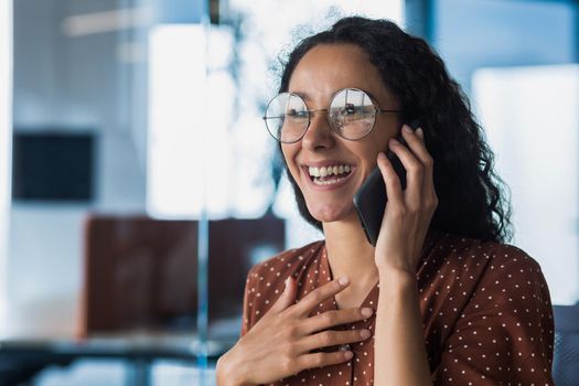 Young and beautiful hispanic woman talking on the phone and smiling happy, business woman close up in glasses and curly hair working inside a modern office building