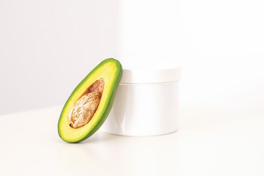 Skincare with natural cosmetics. White cosmetic jar of cream with half of the avocado near against white background, copy space, mockup