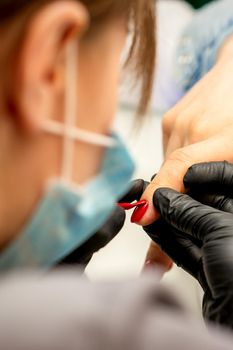 Manicure varnish painting. Close-up of a manicure master wearing rubber black gloves applying red varnish on a female fingernail in the beauty salon