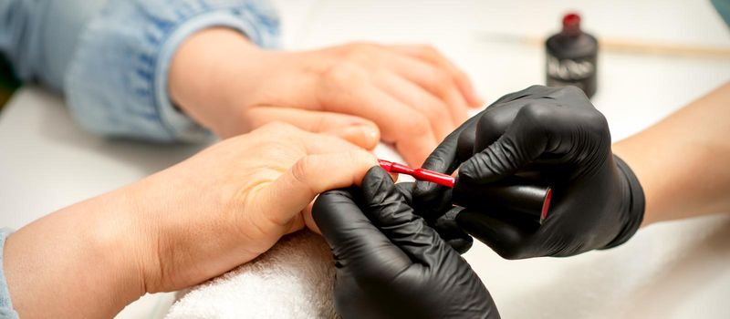 Manicure varnish painting. Close-up of a manicure master wearing rubber black gloves applying red varnish on a female fingernail in the beauty salon