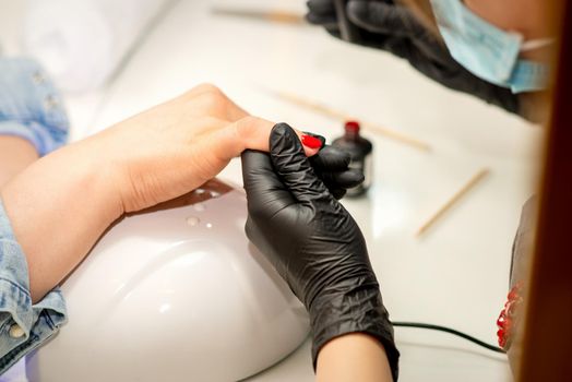 Manicure varnish painting. Close-up of a manicure master wearing rubber black gloves and a protective mask applying red varnish on a female fingernail in the beauty salon