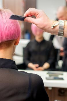 A hairdresser is combing the dyed pink short hair of the female client in hairdresser salon, back view