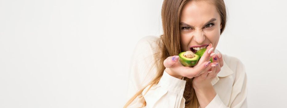 Portrait of a lovely smiling young brunette caucasian woman wearing the white shirt with long hair holding and showing avocado, standing isolated over white background