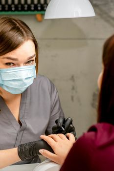 Portrait of young caucasian manicure master wearing protective mask painting female nails in a beauty salon