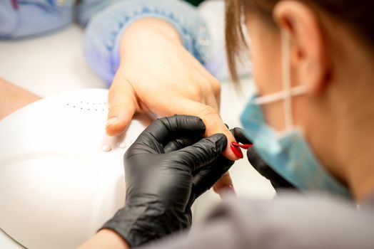 Manicure varnish painting. Close-up of a manicure master wearing rubber black gloves applying red varnish on a female fingernail in the beauty salon