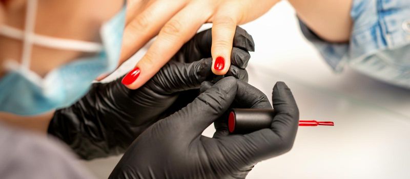 Manicure varnish painting. Close-up of a manicure master wearing rubber black gloves applying red varnish on a female fingernail in the beauty salon