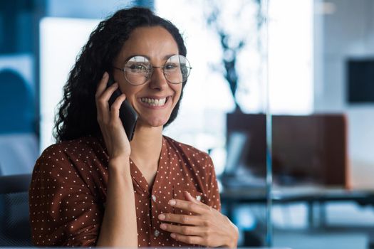 Young and beautiful hispanic woman talking on the phone and smiling happy, business woman close up in glasses and curly hair working inside a modern office building