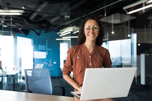 Portrait of young hispanic woman at home, businesswoman smiling and looking at camera waving greeting gesture, freelancer in kitchen using laptop for remote work.