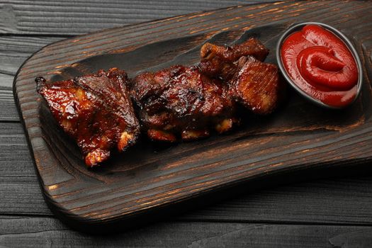 Close up portion of barbecue spare rib slices and BBQ tomato sauce served on wooden board over black table planks, high angle view, personal perspective