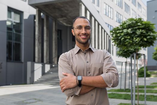 Portrait of young hispanic student man smiling and looking at camera near university campus with arms crossed wearing glasses and shirt.