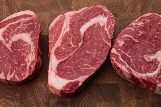 Close up three raw marbled ribeye beef steaks over end grain cutting board of wooden butcher block, high angle view, directly above