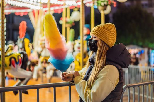 Young woman holding a cotton candy and smiling at a Christmas fair wearing a yellow wool cap wears a medical mask against coronavirus COVID 19.