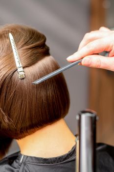 Hairstylist is straightening short hair of young brunette woman with a flat iron in a hairdresser salon, close up