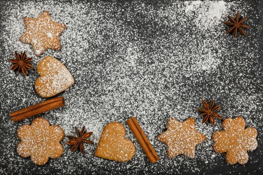 Close up heart and star shaped Christmas gingerbread cookies with cinnamon and star anise spices on black slate background with powdered white sugar icing, elevated top view, directly above