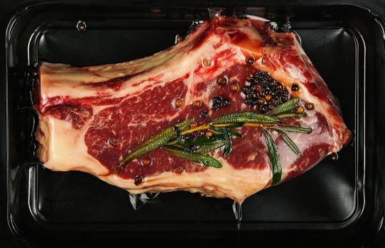 Close up one marbled raw ribeye beef steak with rib bone vacuum sealed with black plastic film, elevated top view, directly above