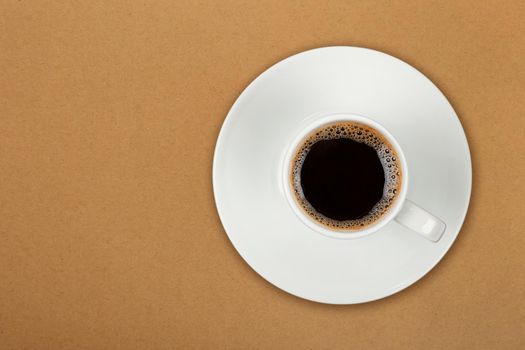 Close up one white cup full of black coffee on saucer over brown paper parchment background, elevated top view, directly above