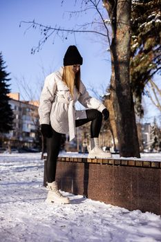 A beautiful young woman in a white jacket and white leather boots walks in a snowy park